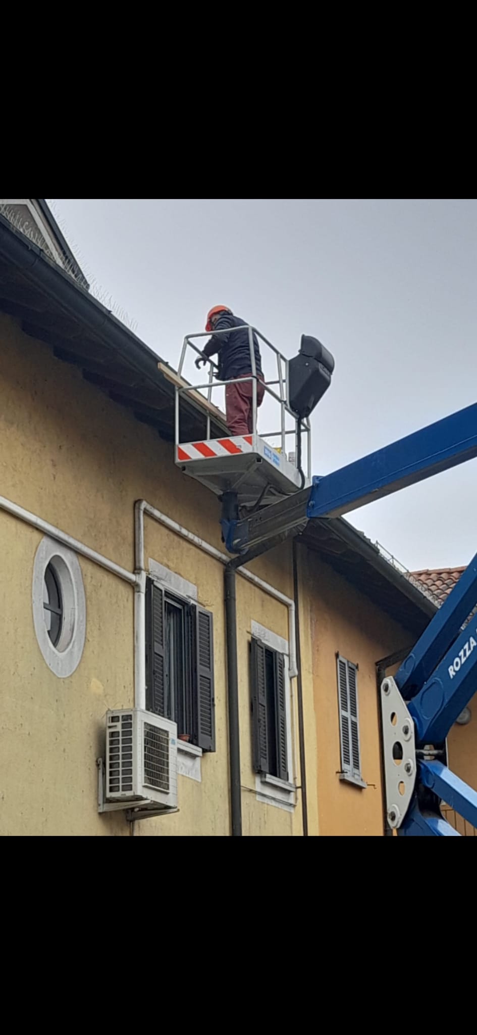 A worker in an elevated platform works beside a building roof and wall-mounted satellite dish.