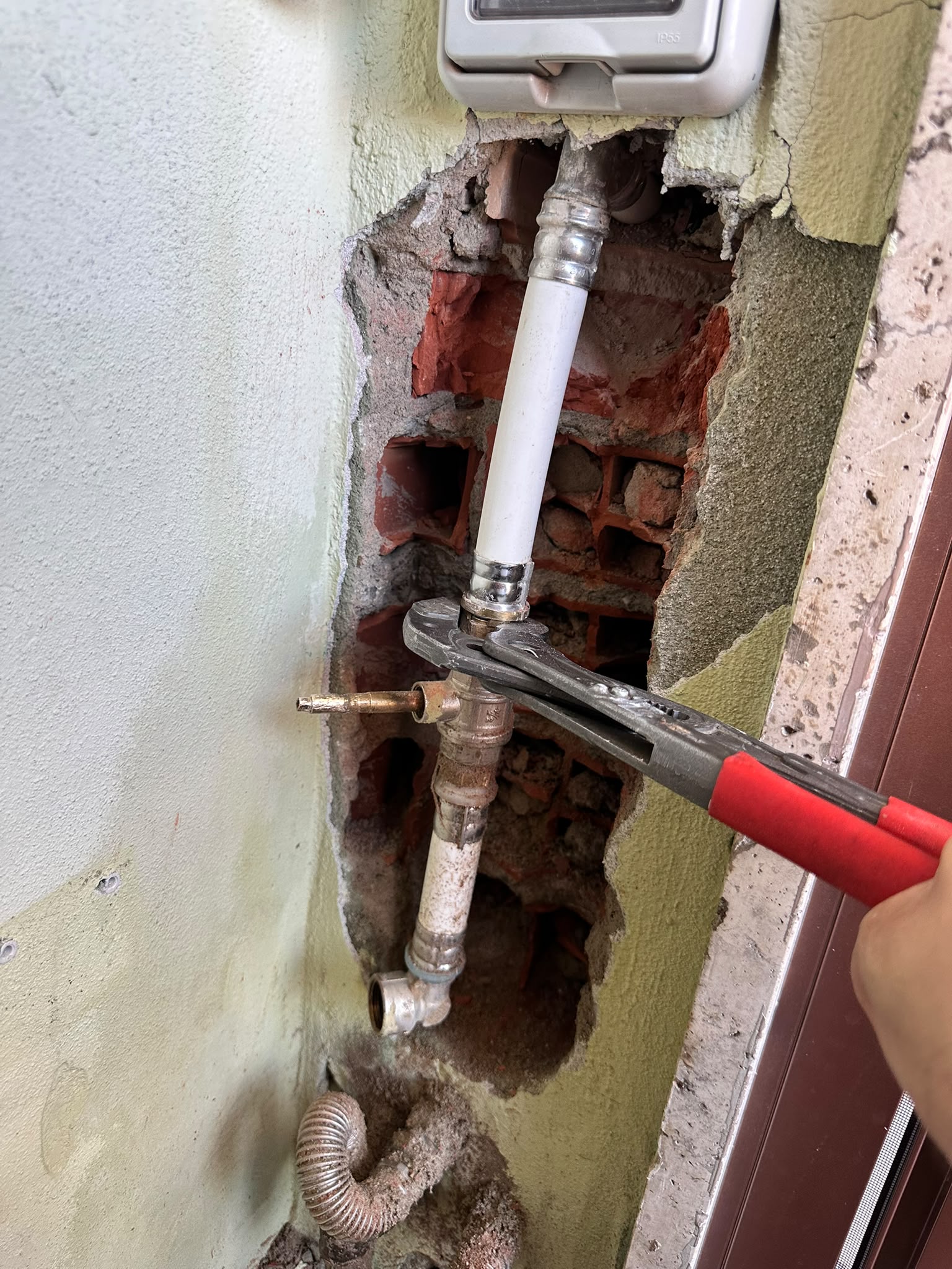 A hand uses red-handled pliers on a pipe fitting inside an exposed wall cavity.